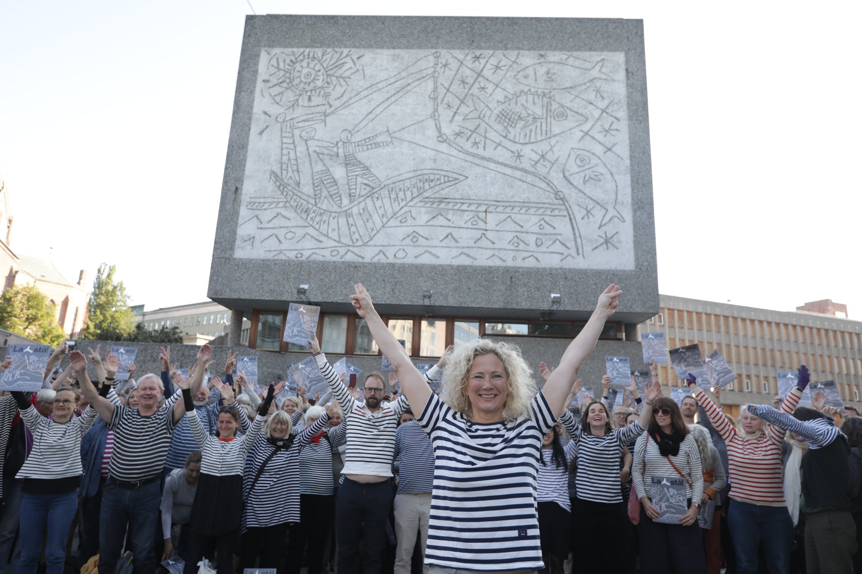 Foto av gavlveggen til Y-blokka med demonstranter med stripete genser foran. I front, Henie Onstad kunstsenters direktør Tone Hansen. Alle med hendene i været (Y-form)