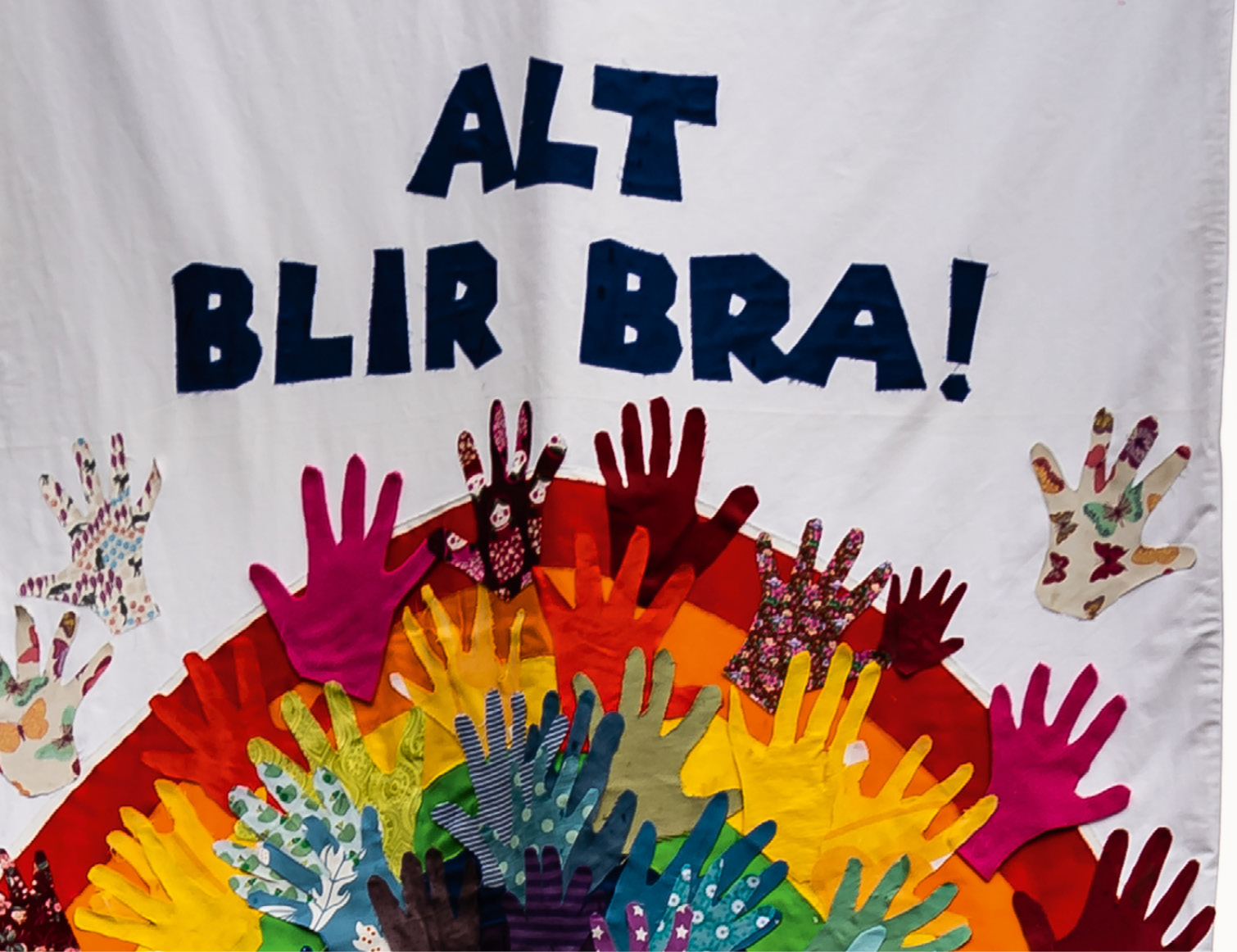 Utsnitt av fane med teksten "Alt blir bra", regnbuemotiv dekorert med barnehender klipt ut av stoff
