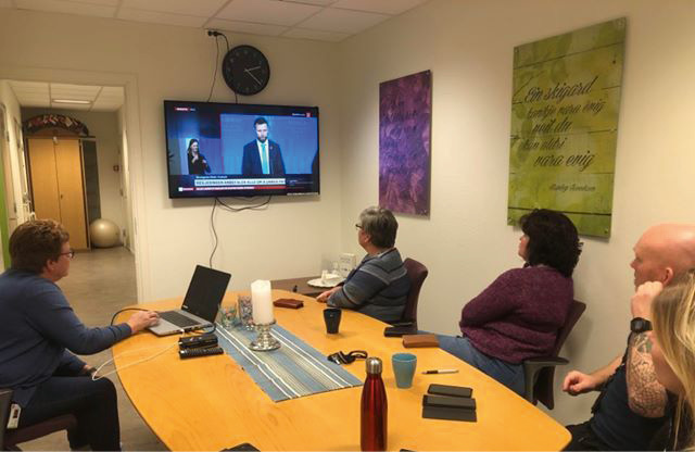 Fire personer rundt et møtebord ser på tv-skjerm som viser pressekonferanse