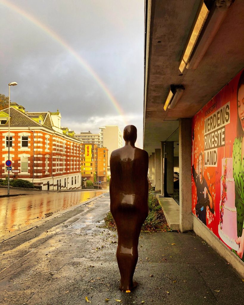 Gatebilde med skulptur i forgrunnen og regnbue i bakgrunnen